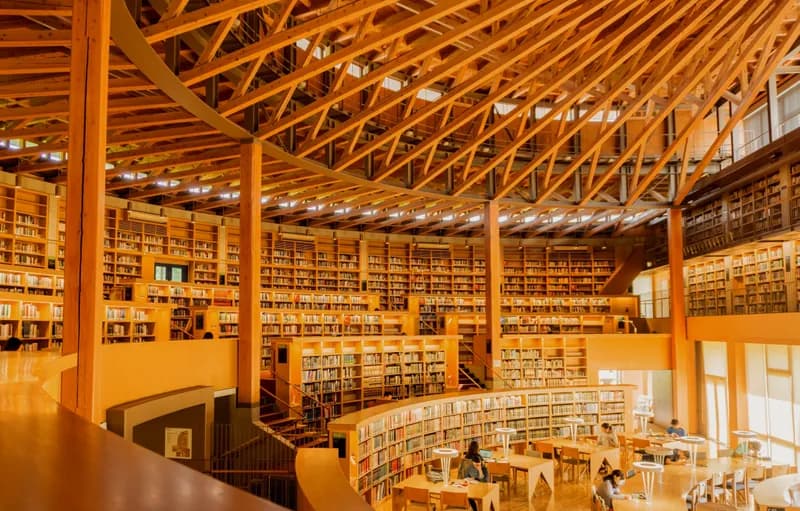 Modern library interior with exposed wooden beam ceiling and multi-level bookshelves filled with books, featuring reading areas with tables and visitors studying throughout the space