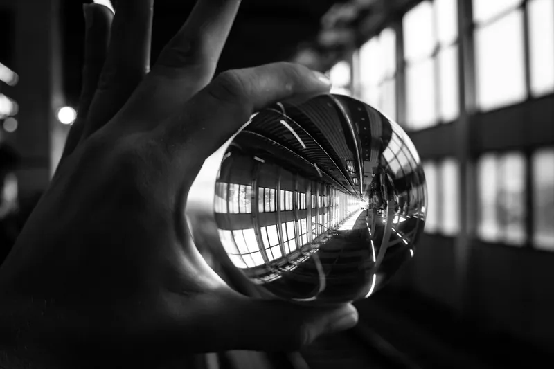 Photograph of a hand holding a glass sphere that refracts a long windowed corridor, creating a miniature inverted world inside the ball