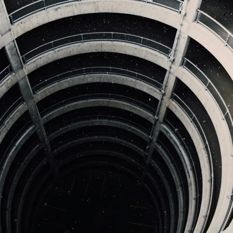 Picture looking down a winding parking-lot ramp many floors deep. The bottom is distant, dark, and gloomy. It's snowing.