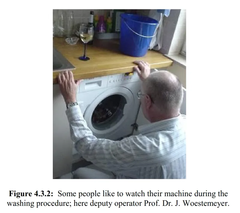 Picture of a man kneeling in front of a washing machine and looking at it. There is a wine glass on the machine.