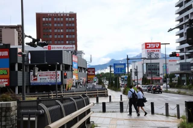 A rainy day in a Japanese commercial district. Two students in school uniforms cross a wet pedestrian area near a road intersection. A brown "UNIZO INN Express" hotel stands on the left, while a "コジマ" (Kojima) electronics store sign is visible on the right. Mountains are faintly visible through clouds in the background.