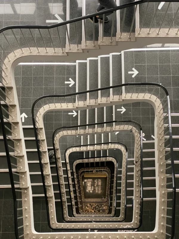 View down a square stairwell. Flights of stairs with black handrails spiral down to the ground floor, and white arrows on the floor tiles show the way.