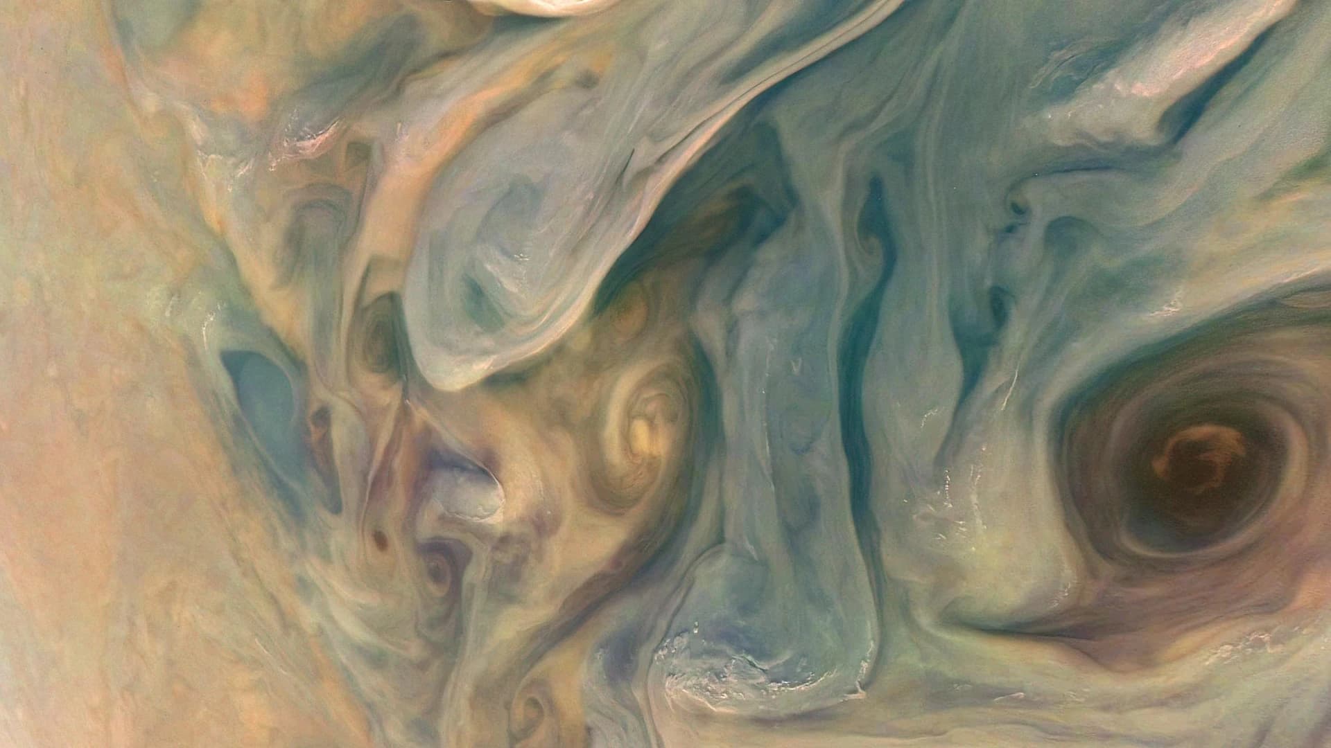 Close-up of swirling clouds on Jupiter in pale blue, cream, and orange, with a dark brown vortex on the right.