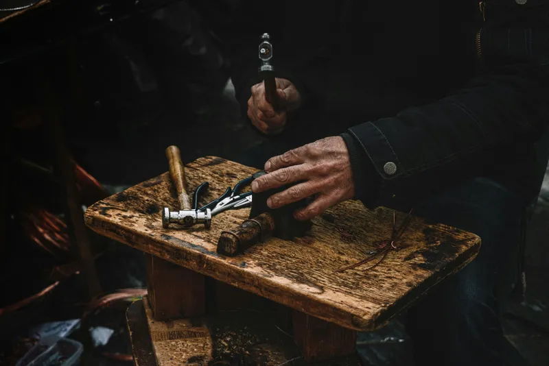 An artisan's weathered hand rests on a worn wooden bench holding small hammers and pliers, while the other hand raises a hammer.