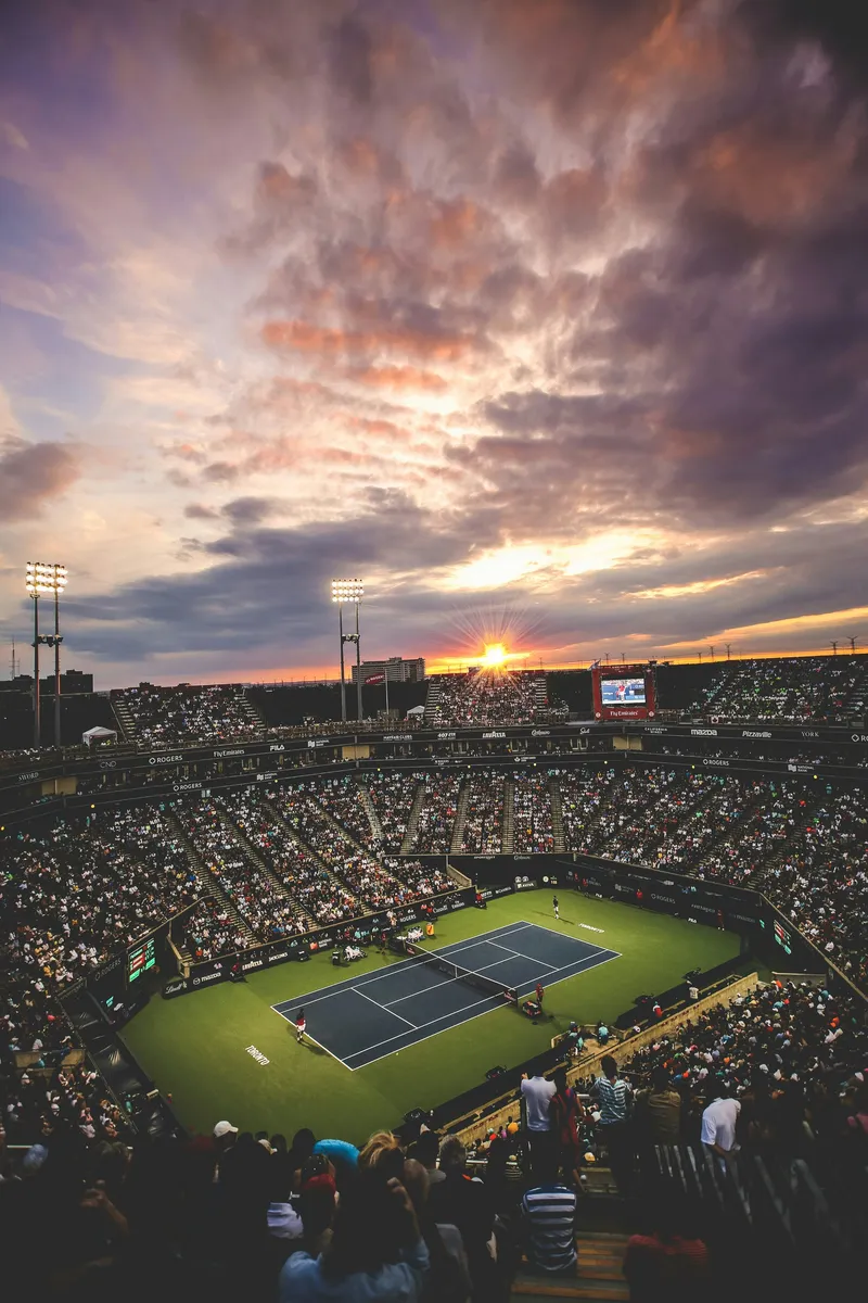A panoramic view of a tennis stadium at sunset, taken from elevated seating. The image shows a packed tennis venue with thousands of spectators filling tiered seating sections surrounding a blue tennis court. Above, a dramatic sky displays rich orange, pink, and purple clouds illuminated by the setting sun, which appears as a bright starburst on the horizon. Stadium floodlights are visible on tall poles, and the scene captures the grand scale and atmosphere of a major tennis tournament.