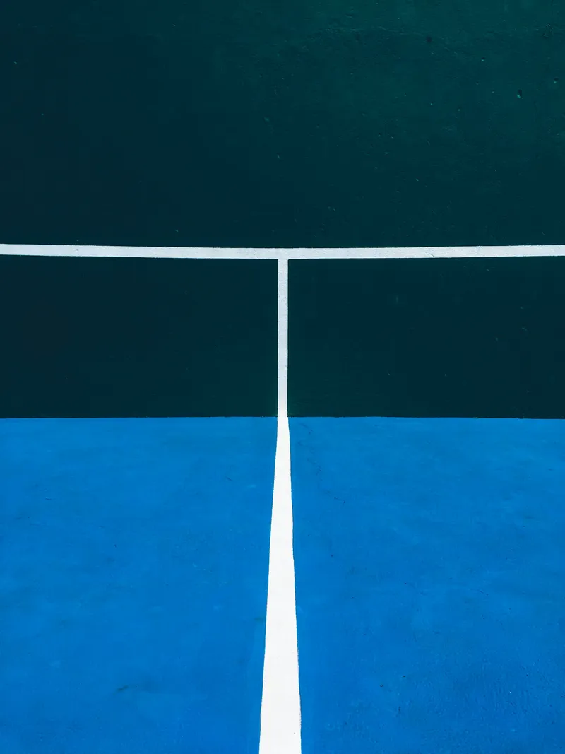 A minimalist geometric composition showing a tennis court from above, divided into sections by white court lines. The image features a stark contrast between dark teal-green and bright blue court surfaces, with crisp white lines creating a grid pattern. The clean, abstract composition emphasizes the geometric precision of tennis court design, with the different colored surfaces creating distinct rectangular sections.