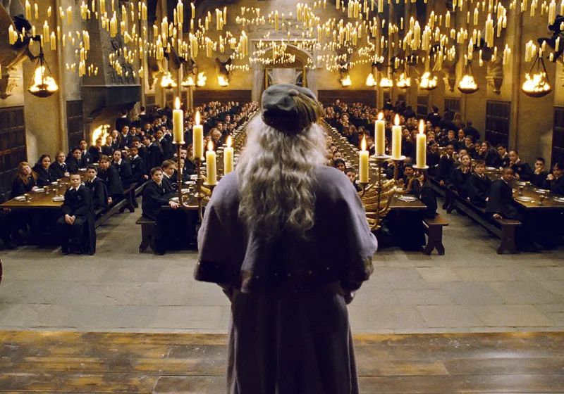 A robed figure stands facing rows of seated students at long tables in a grand hall lit by hundreds of floating candles.