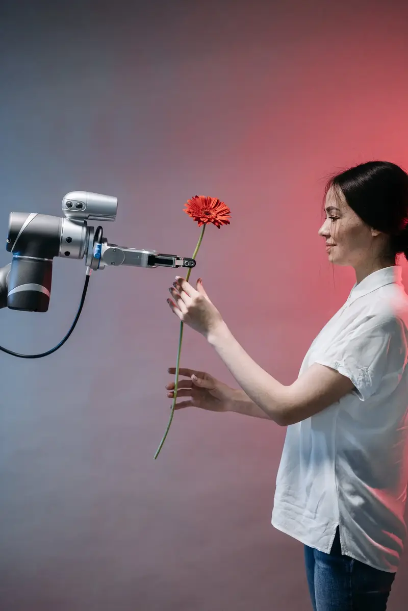 A robotic arm holds out a red gerbera flower to a smiling woman in a white shirt, who reaches out to take it.