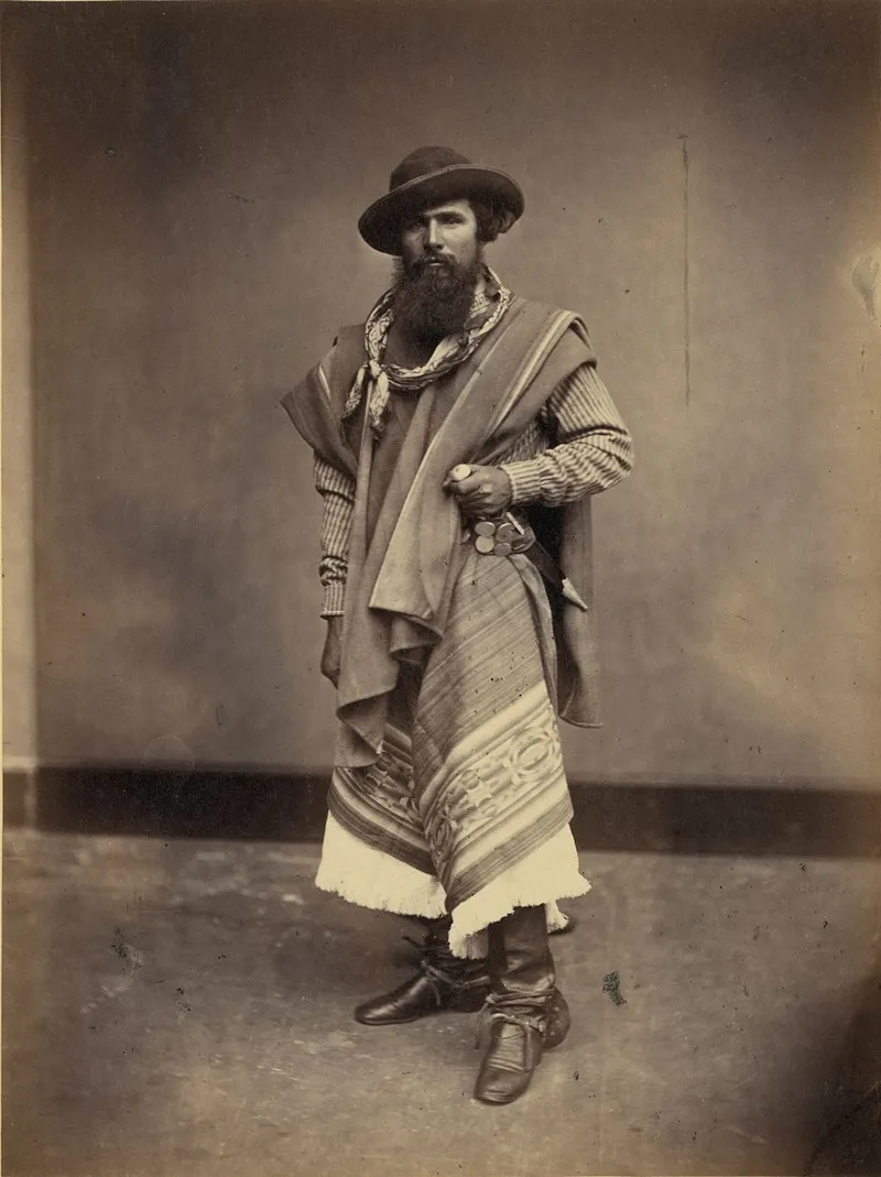 Sepia photograph of a bearded gaucho in traditional dress: wide-brimmed hat, poncho draped over shoulders, striped chiripá, and leather boots, with a decorative coin-studded belt and knife at his waist