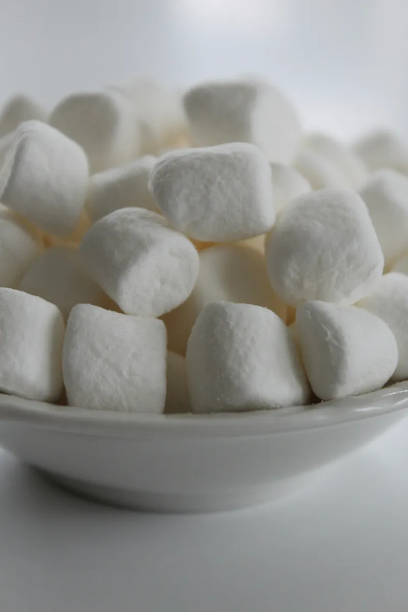 A close-up photograph of white marshmallows piled in a white ceramic bowl. The marshmallows are soft, pillowy, and cylindrical in shape with rounded edges, creating gentle shadows between them.