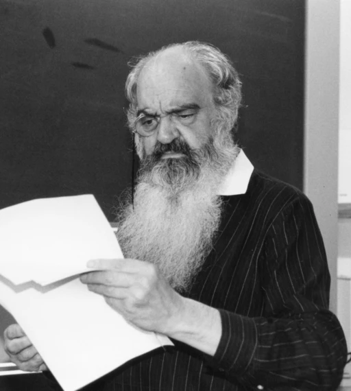 Black and white photograph of Stafford Beer with a long white beard and a monocle, reading papers