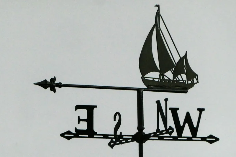 An iron weather vane with the shape of a sailboat riding an arrow, and the four cardinal directions N, S, E, W.