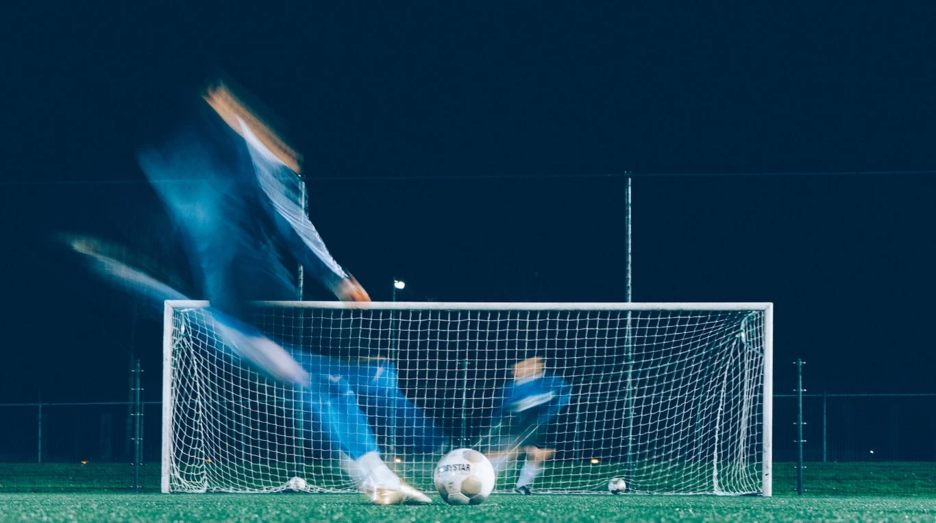 A soccer player takes a penalty kick as the goalkeeper dives across the goal.