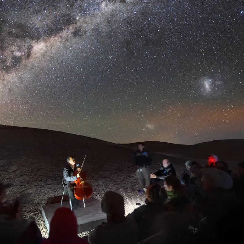 Picture of the cellist Yo-Yo Ma sitting in front of a small crowd under a starry sky.